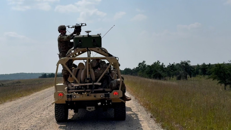 A soldier operates a drone from the back of a military vehicle on a dirt road in a field.