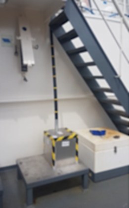 Industrial room with a dark metal staircase, a silver box with hazard tape, a striped pole, and a white container.