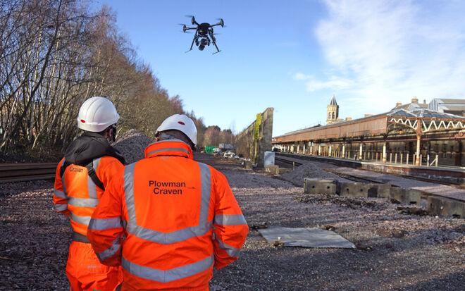 Two workers in orange safety vests watch a drone fly over a railway line construction site.